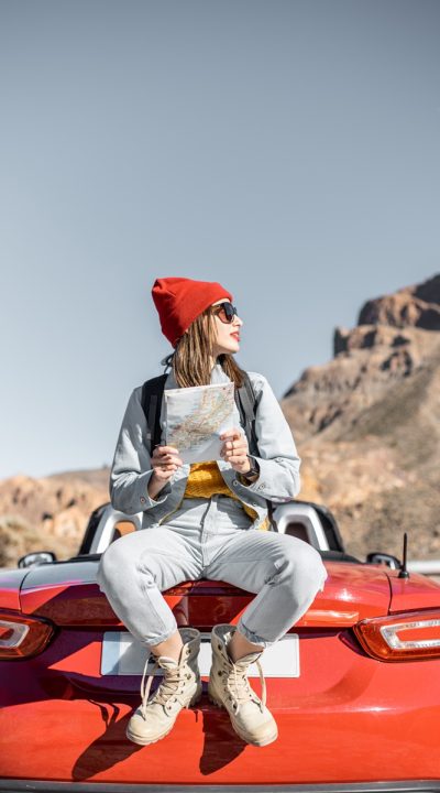 Woman traveling by car on the dessert valley Lifestyle portrait of a carefree woman dressed casually in jeans and red hat sitting with map on the car trunk, enjoying road trip on the desert valley